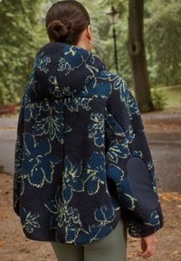 Granatowa bluza polarowa z kapturem, ozdobiona kwiatowym wzorem, wykonana z fakturowanej tkaniny, o luźnym kroju i ciemnoniebieskich łatach na łokciach.