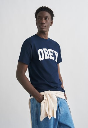 Νέος άνδρας που φοράει μπλε σκούρο μπλουζάκι με την επιγραφή "OBEY", ανοιχτόχρωμα τζιν και μπεζ φούτερ δεμένο στη μέση, κοιτώντας προς το πλάι.