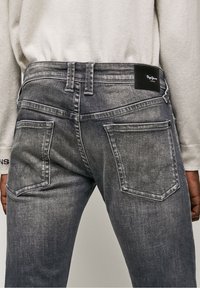 Jeans de mezclilla gris con un acabado desgastado y texturizado. Presenta una etiqueta de cuero negro y dos bolsillos traseros con costuras curvadas. Incluye lazos en la cintura.