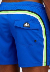 Pantaloni da bagno blu con tasca laterale, strisce di accento verdi, nere e bianche, e una piccola etichetta logo vicino alla tasca posteriore.