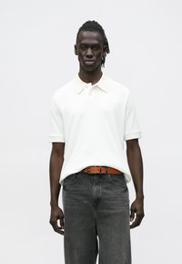 Mladý muž s tmavou pokožkou a copánky, oblečený v bílé krátkosvlé polokošili, hnědém pásku a tmavě šedých džínech, stojící před bílým pozadím.