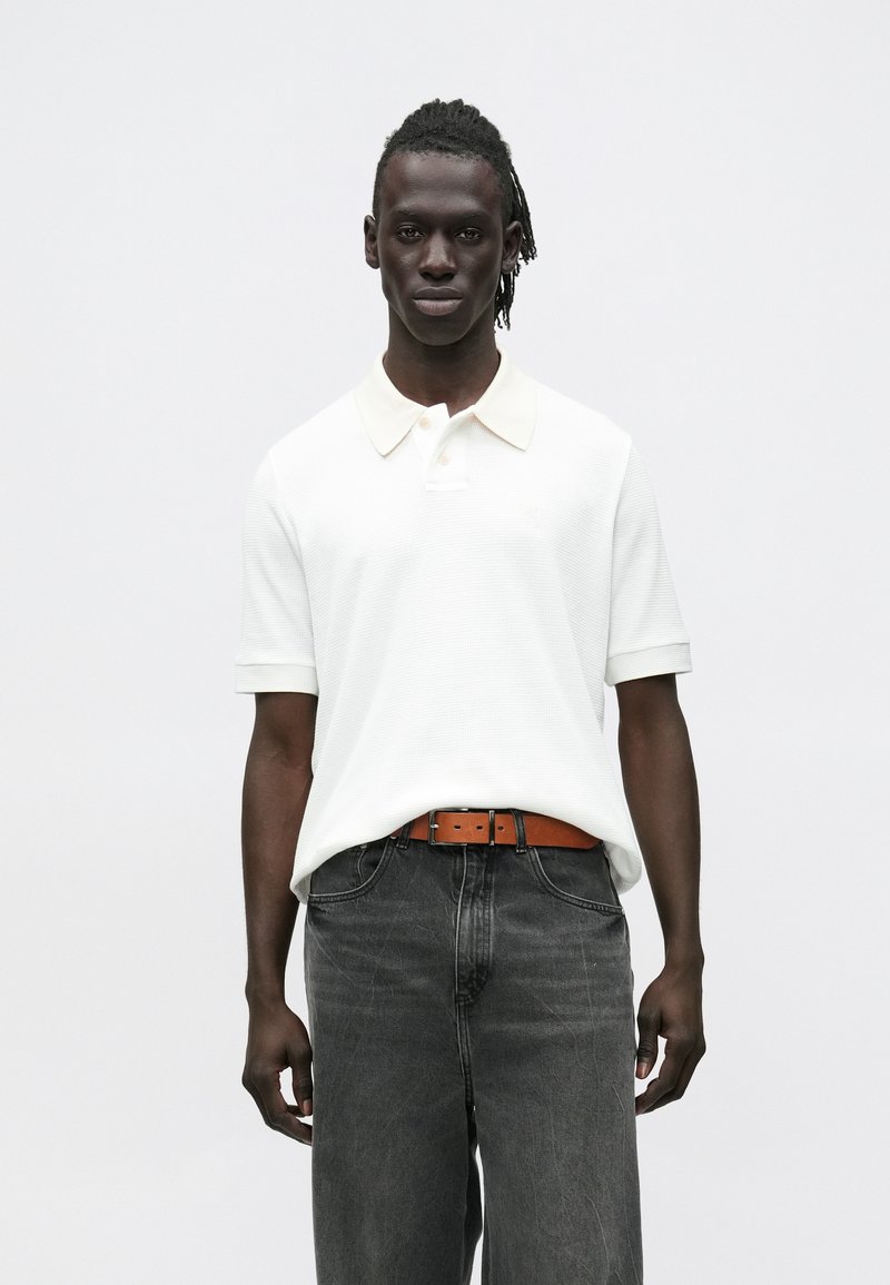 Mladý muž s tmavou pokožkou a copánky, oblečený v bílé krátkosvlé polokošili, hnědém pásku a tmavě šedých džínech, stojící před bílým pozadím.