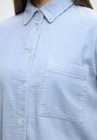 Svetlomodrá košeľa na zapínanie s textúrou, golierom, vertikálnou rebrovou tkaninou, predným vreckom a bielymi gombíkmi.