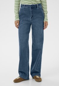 Jeans de denim de pierna ancha en azul oscuro con una textura sutil. Presenta una cintura alta, pasadores para cinturón y bolsillos frontales. Combinados con zapatos marrones.