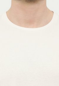 Κοντινό πλάνο του λαιμού και του πάνω μέρους του στήθους ενός ατόμου που φοράει μια απλή λευκή μπλούζα με στρογγυλή λαιμόκοψη, σε λευκό φόντο.