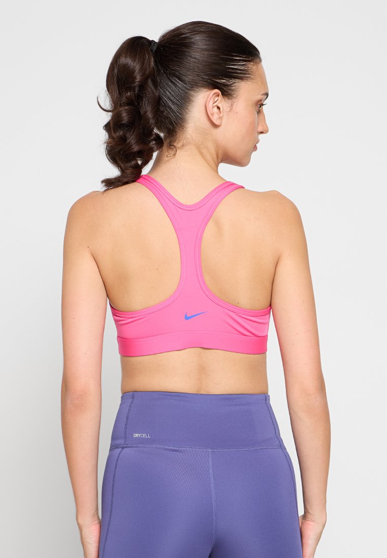 Mujer con coleta rizada que lleva un sujetador deportivo rosa de Nike y leggings morados de talle alto con el texto "DRYCELL" en la cadera izquierda, mostrada desde atrás.