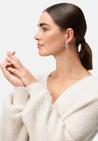 Silberne Creolen mit einem geschichteten Design, getragen von einer Person mit glattem Haar, in einem weichen beigen Pullover mit V-Ausschnitt und Ripptextur.