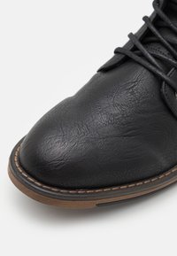 Černá kožená bota s texturovaným povrchem, kulatou špičkou, šněrováním a prošívanou hnědou podrážkou.