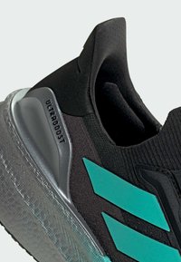 Black athletic shoe with a textured upper, teal accents, and a transparent rubber sole featuring "ULTRAGBOOST" branding on the heel.