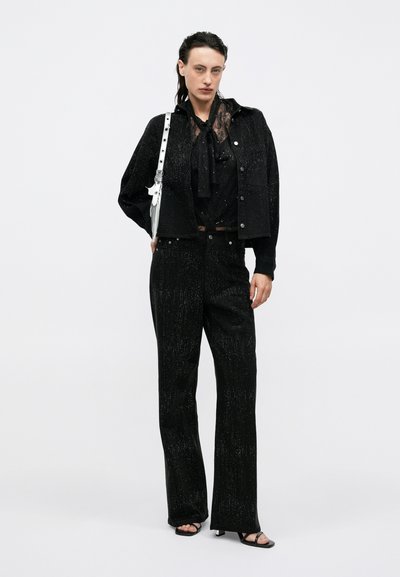 Outfit nero con un blazer testurizzato e scintillante, una blusa con colletto a laccio e pantaloni coordinati. Include una borsa bianca e décolleté nere aperte.
