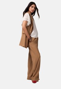 Gilet beige senza maniche sovrapposto a una maglietta bianca con maniche in pizzo, abbinato a pantaloni larghi beige e scarpe rosse, che mostrano una texture liscia.