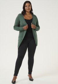 Groene gebreide cardigan met kabelpatroon, open voorkant en lange mouwen. Draagt over een zwarte top en gecombineerd met zwarte skinny jeans.