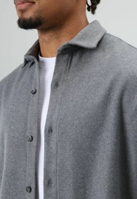 Grijze wollen overhemd met knopen en een kraag, met een zachte textuur en zwarte knopen, gedragen over een wit T-shirt. Close-up weergave.