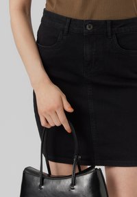 Jupe en denim noire avec poches avant, design ajusté et fermeture à boutons, associée à un sac à main noir avec une texture lisse et une forme structurée.