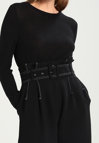 Top negro de mangas largas combinado con pantalones de tiro alto y pernera ancha, que cuentan con un cinturón audaz y costuras blancas en los bolsillos y las costuras.