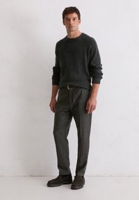 Dunkelgrauer Strickpullover mit gerippten Bündchen, kombiniert mit grauen Nadelstreifenhosen. Das Modell trägt braune Slipper und steht vor einem neutralen Hintergrund.