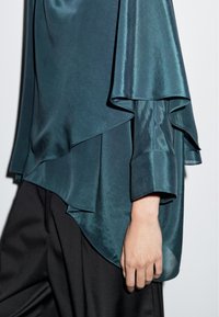Satinblus i teal med lager-på-lager design, förlängda ärmar och en flödande silhuett, kombinerad med svarta byxor.
