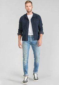 Marineblauwe spijkerjas met knopen aan de voorkant, twee borstzakken, wit T-shirt, lichtblauwe versleten skinny jeans en zwart-witte sneakers.