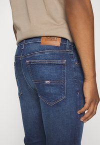 Tmavě modré džíny z denimu s koženou nášivkou, kontrastním šitím a dvěma zadními kapsami, přičemž jedna z nich má malý detail loga značky.