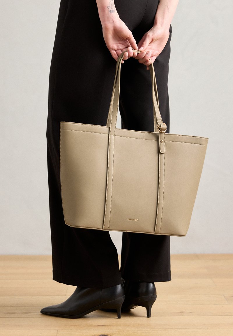 Bolsa tote de couro bege com duas alças longas, forma estruturada, costura mínima e detalhe de logotipo na frente. Usada com roupa preta.