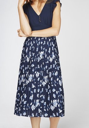 Nainen pukeutuneena tummansiniseen mekkoon, jossa on napillinen V-kaula-aukko ja laskostettu kukkakuvioinen hame, seisomassa käsivarret ristissä ja käsi koskettaa kasvoja.