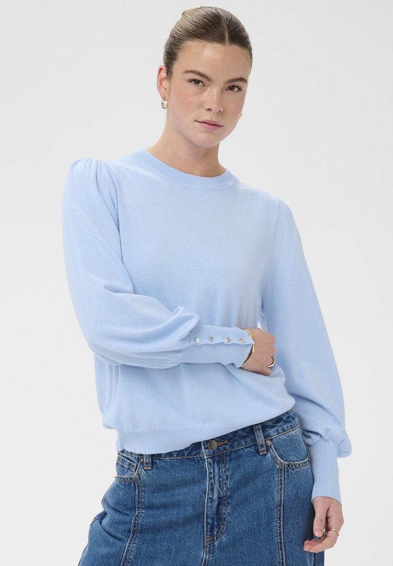 Pull bleu clair avec manches bouffantes et poignets boutonnés, fabriqué dans un matériau doux. Associé à un jean taille haute en denim.