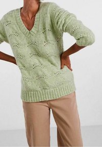 Pull tricoté vert clair avec un col en V, présentant un motif en dentelle et des poignets et ourlet côtelés. Porté avec un pantalon beige.