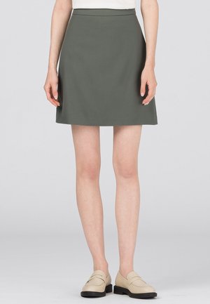 Kobieta w oliwkowo-zielonej spódnicy do kolan, białej bluzce i beżowych mokasynach, stojąca na tle jednolitego, jasnego tła.