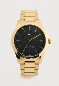 Gold metal wristwatch with black dial, gold hour markers, three gold hands, and a small rectangular red, white, and blue logo above "HILFIGER" text.