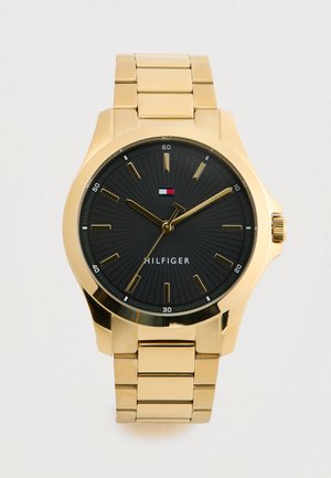 Gold metal wristwatch with black dial, gold hour markers, three gold hands, and a small rectangular red, white, and blue logo above "HILFIGER" text.