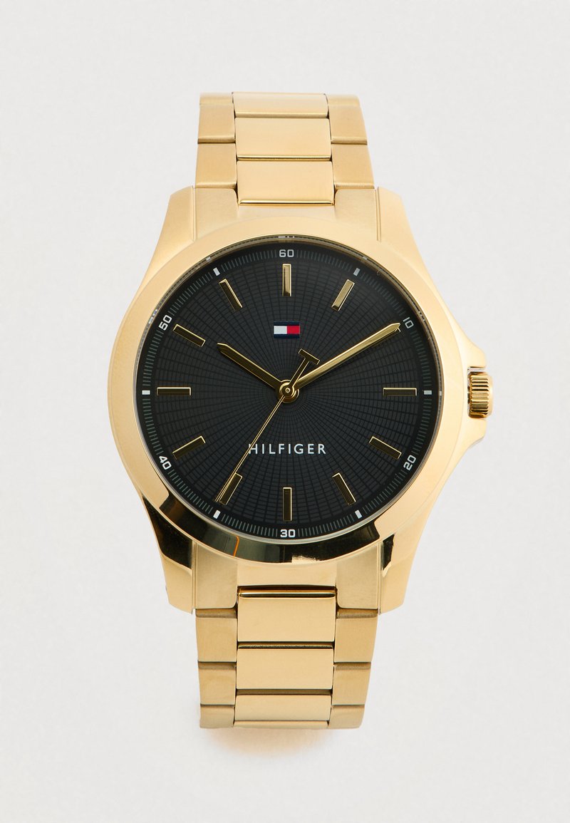 Gold metal wristwatch with black dial, gold hour markers, three gold hands, and a small rectangular red, white, and blue logo above "HILFIGER" text.