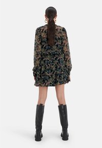 Schwarz mit Blumenmuster versehenes Kleid mit langen Ärmeln und Rüschensaum. Das Model trägt hohe schwarze Stiefel. Der Stoff hat ein transparentes Overlay.