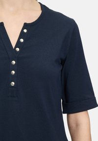 Granatowa koszulka polo z dekoltem w serek, podwiniętymi rękawami i srebrnymi zatrzaskami wzdłuż listwy. Wykonana z miękkiego, lekkiego materiału.