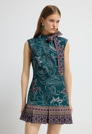 Robe verte mi-longue avec un motif paisley, décolleté rond et détail de nœud sur le côté. L'ourlet évasé présente des accents géométriques contrastants.