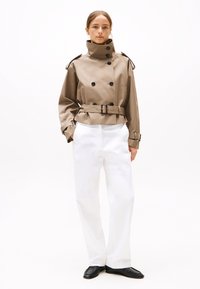 Trench-coat marron clair, résistant à l'eau, avec un col montant, des boutons double boutonnage et une taille ceinturée, associé à un pantalon large blanc.