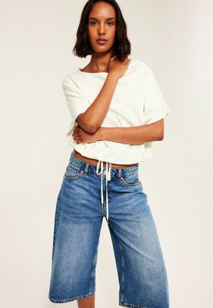 Kvinde iført en hvid kortærmet top og vidde blå denimshorts, står med den ene hånd på nakken og den anden krydset over kroppen.