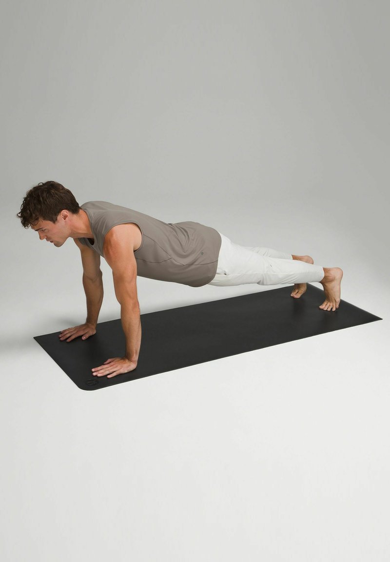 Černá jóga podložka s osobou v pozici prkna, oblečenou ve šedém tílku bez rukávů a bílých kalhotách, která ukazuje svalový tonus a správné zarovnání.