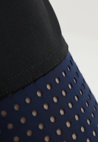 Musta vyö siirtyy tummaan siniseen kangasta, jossa on tasaisesti sijoitettuja rei'ityksiä, luoden teksturoituneen ja hengittävän muotoilun.