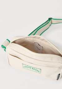 Μπεζ καμβά τσάντα ώμου με πράσινο ριγέ λουρί, διαθέτει μπροστινή τσέπη και κλείσιμο με φερμουάρ. Περιλαμβάνει εσωτερική ετικέτα με λογότυπο.