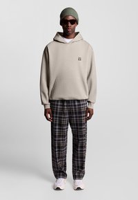 Felpa con cappuccio beige e tasca frontale, abbinata a pantaloni a quadri in blu marino, giallo e bianco. Gli accessori includono un berretto verde chiaro e occhiali da sole.