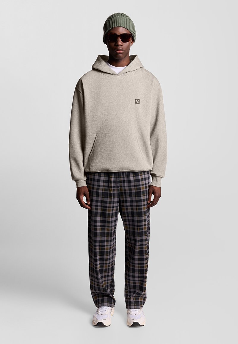 Felpa con cappuccio beige e tasca frontale, abbinata a pantaloni a quadri in blu marino, giallo e bianco. Gli accessori includono un berretto verde chiaro e occhiali da sole.