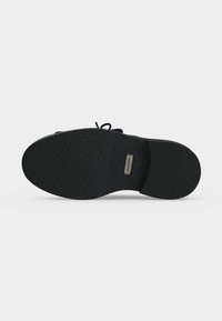 Semelle de chaussure habillée noire avec une prise texturée, une petite étiquette de marque métallique rectangulaire près de la voûte plantaire, et des zones lisses au talon et aux orteils.