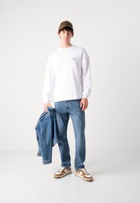 Sudadera blanca con logo azul, jeans de mezclilla azul, zapatillas gris-marrón y una gorra marrón, sosteniendo una chaqueta de mezclilla sobre el brazo.