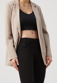 Femme portant un blazer beige, un top court noir, un pantalon noir taille haute et un collier de fleurs coloré sur un fond uni.