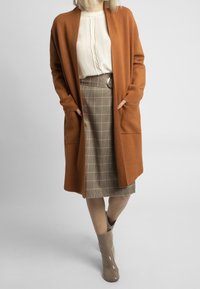 Brązowy długi kardigan, kremowa bluzka, plisowana ołówkowa spódnica w kratę z paskiem oraz beżowe błyszczące botki do kostki. Gładka tkanina i stonowany krój.