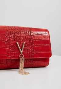 Borsa in pelle rossa con texture a coccodrillo, dotata di una chiusura a forma di V in tono oro e un dettaglio con frangia a catena; forma rettangolare con finitura liscia.