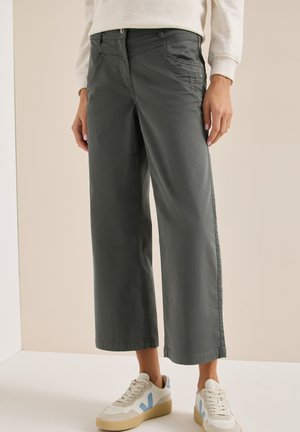 Persona vistiendo pantalones anchos gris oscuro, zapatillas blancas y azules, y una camiseta de manga larga beige clara, de pie contra un fondo sencillo.