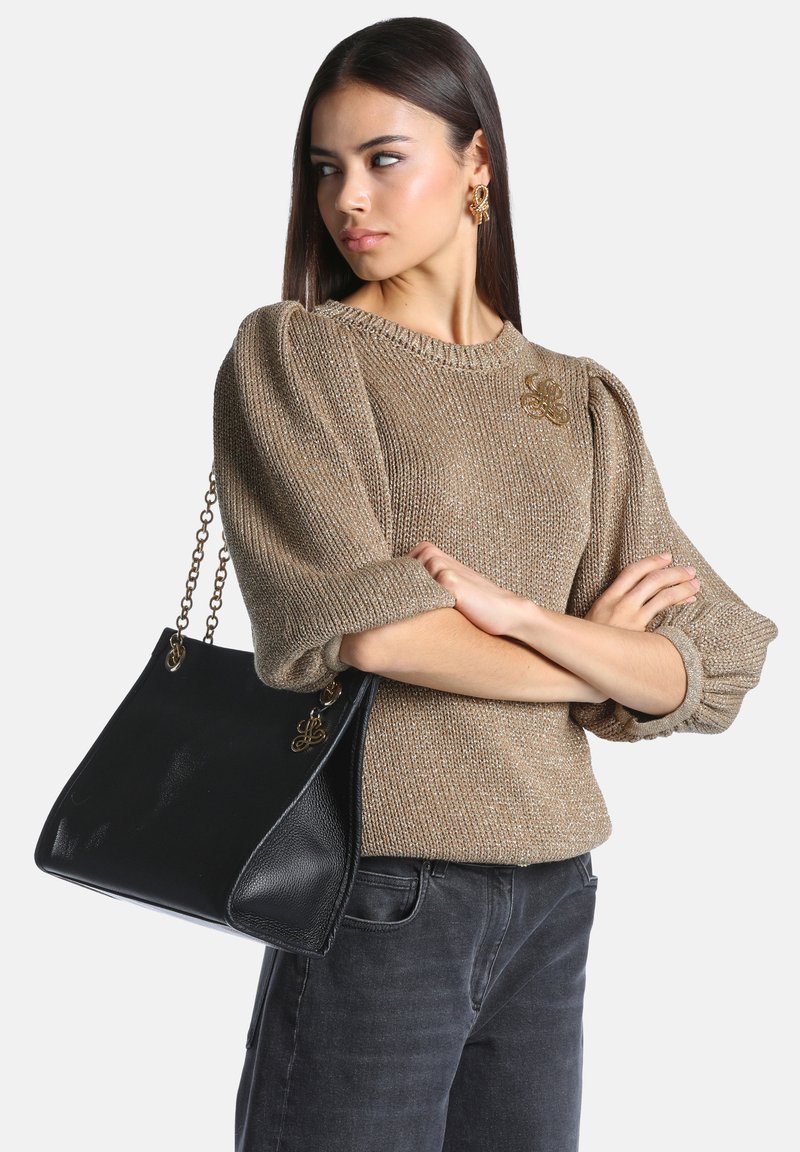 Femme aux longs cheveux noirs portant un pull beige texturé, un jean noir, des boucles d'oreilles dorées et portant un grand sac à main noir avec des anses en chaîne dorée.