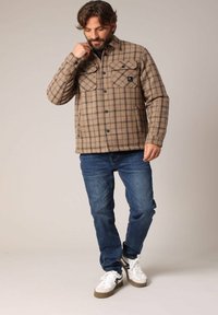 Geverifieerde flanellen shirtjas in bruine tinten met zwarte accenten. Voorzien van voorzakken en knoopsluiting. Geregistreerd met blauwe spijkerbroeken en sneakers.