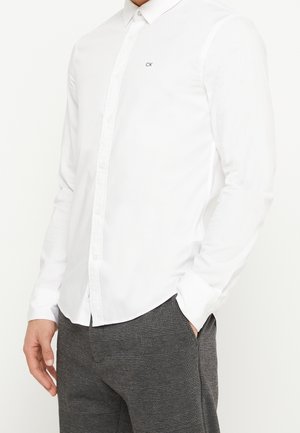 Homme portant une chemise blanche ajustée boutonnée avec un petit logo « CK » sur la poitrine et un pantalon gris à carreaux, une main dans la poche.